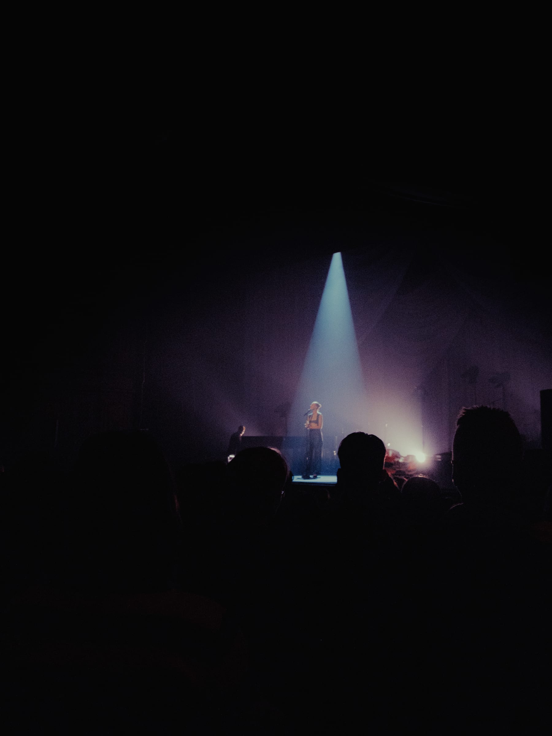 A figure on a far stage under a single beam, heads of the audience in silhouette