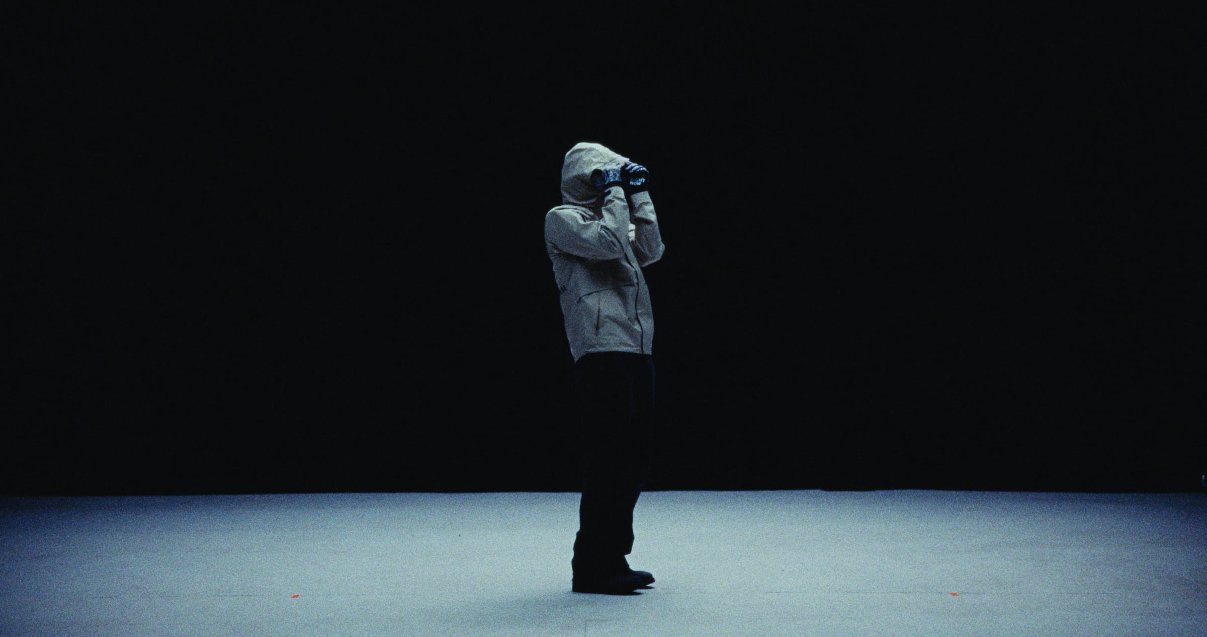 A hooded figure alone on a dark stage, hands raised to their face